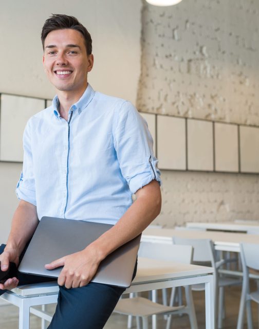 young attractive smiling man sitting in co-working open office, holding laptop, self-employed, freelance job, education online